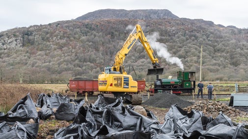 A yellow digger in front of a steam engine, with dozens of black sacks at the front of the picture waiting to be loaded